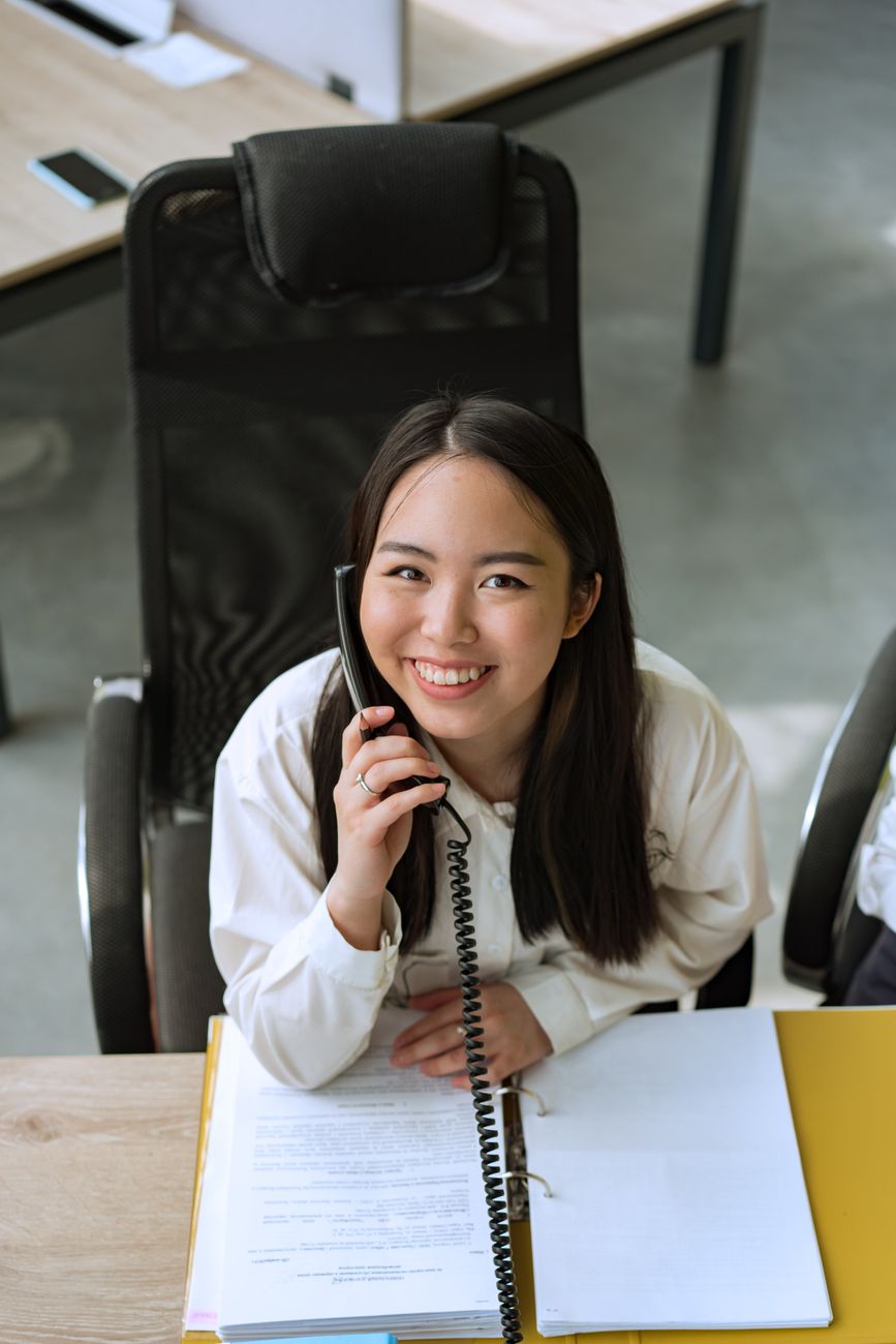 Atendente sorrindo ao telefone em secretaria escolar durante atendimento